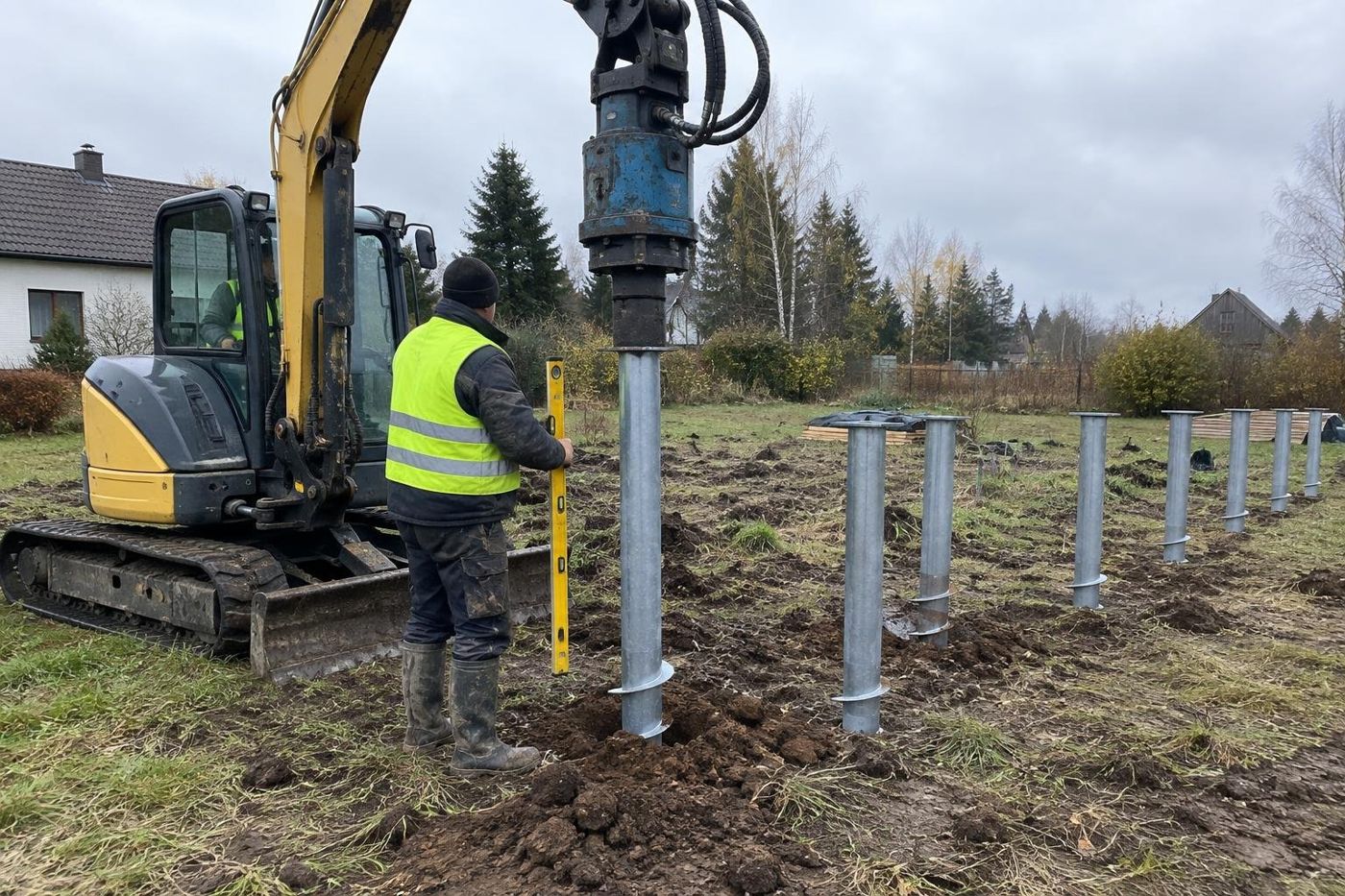 Miniekskavaator keerab terasest kruvivaia maasse, taustal rida juba paigaldatud vaiu